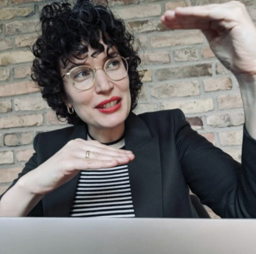 Eine Frau mit Brille sitzt mit einem Laptop an einem Schreibtisch.