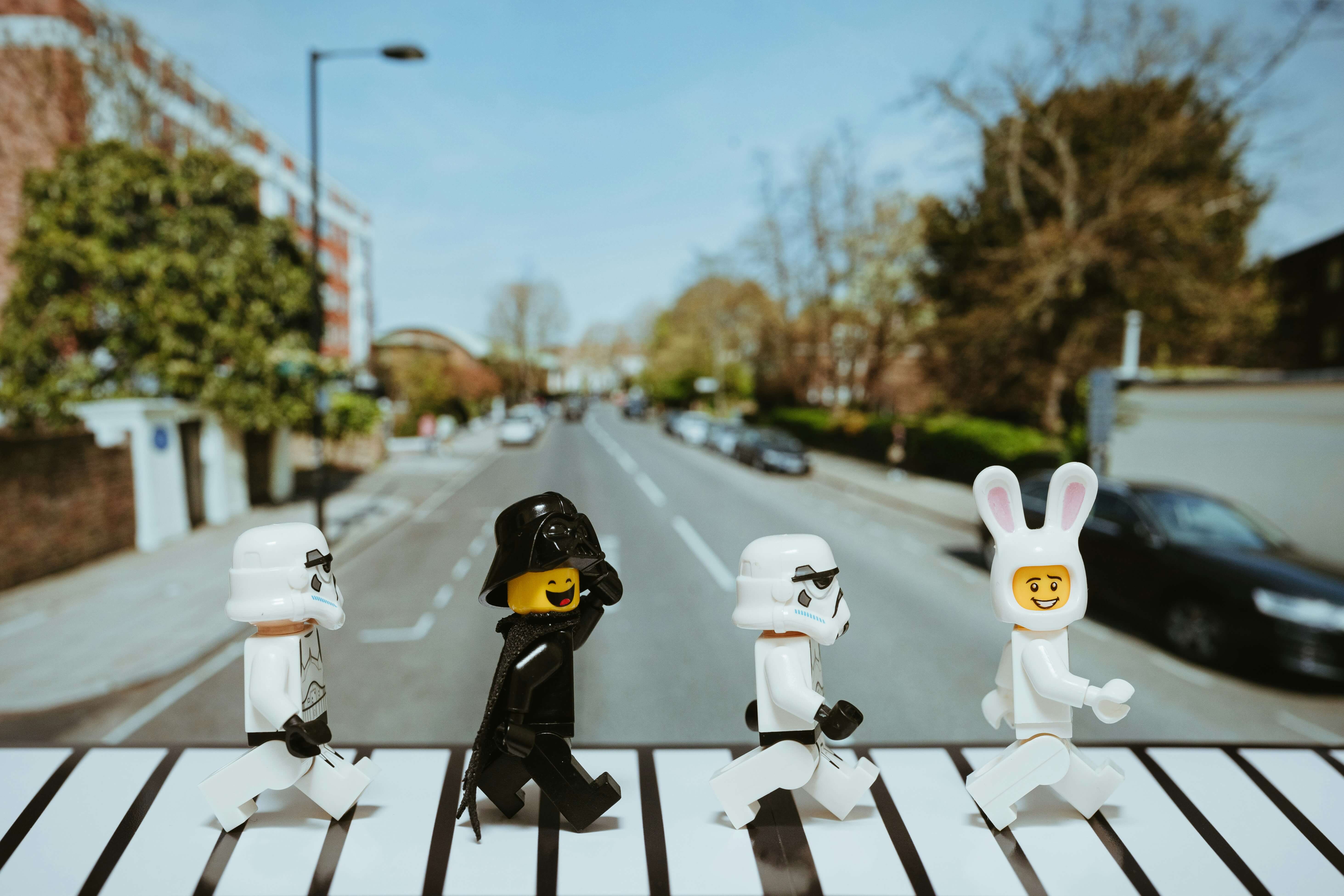 Miniaturfiguren, die an Star Wars-Sturmtruppen und Darth Vader erinnern, wobei eine Figur ein Hasenkostüm trägt, auf einem Zebrastreifen mit einer Straße im Hintergrund.