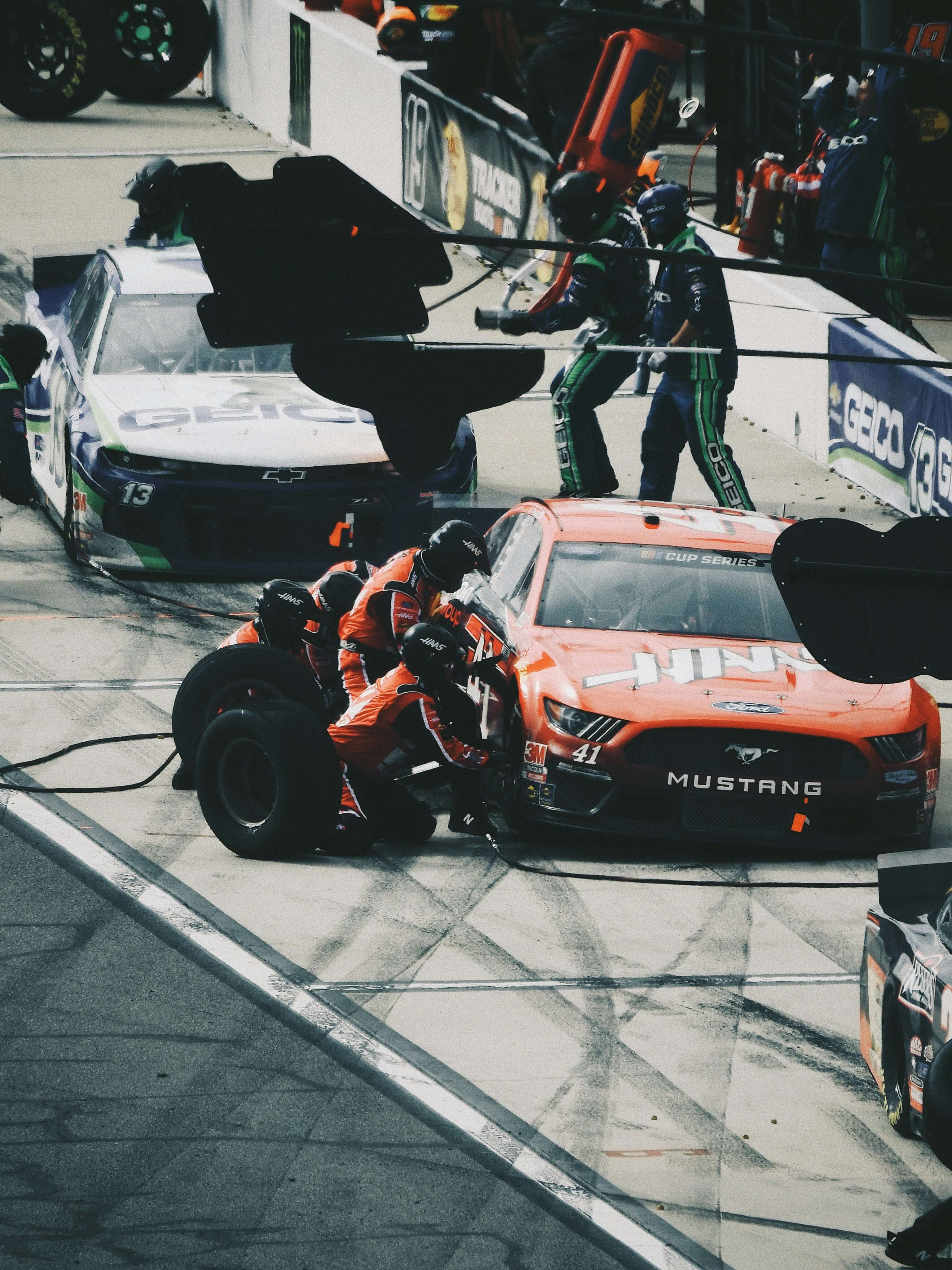 Eine Boxencrew wartet einen orangefarbenen Ford Mustang-Rennwagen, während sich während eines Rennens im Hintergrund ein weiteres Auto bei einem Boxenstopp befindet.