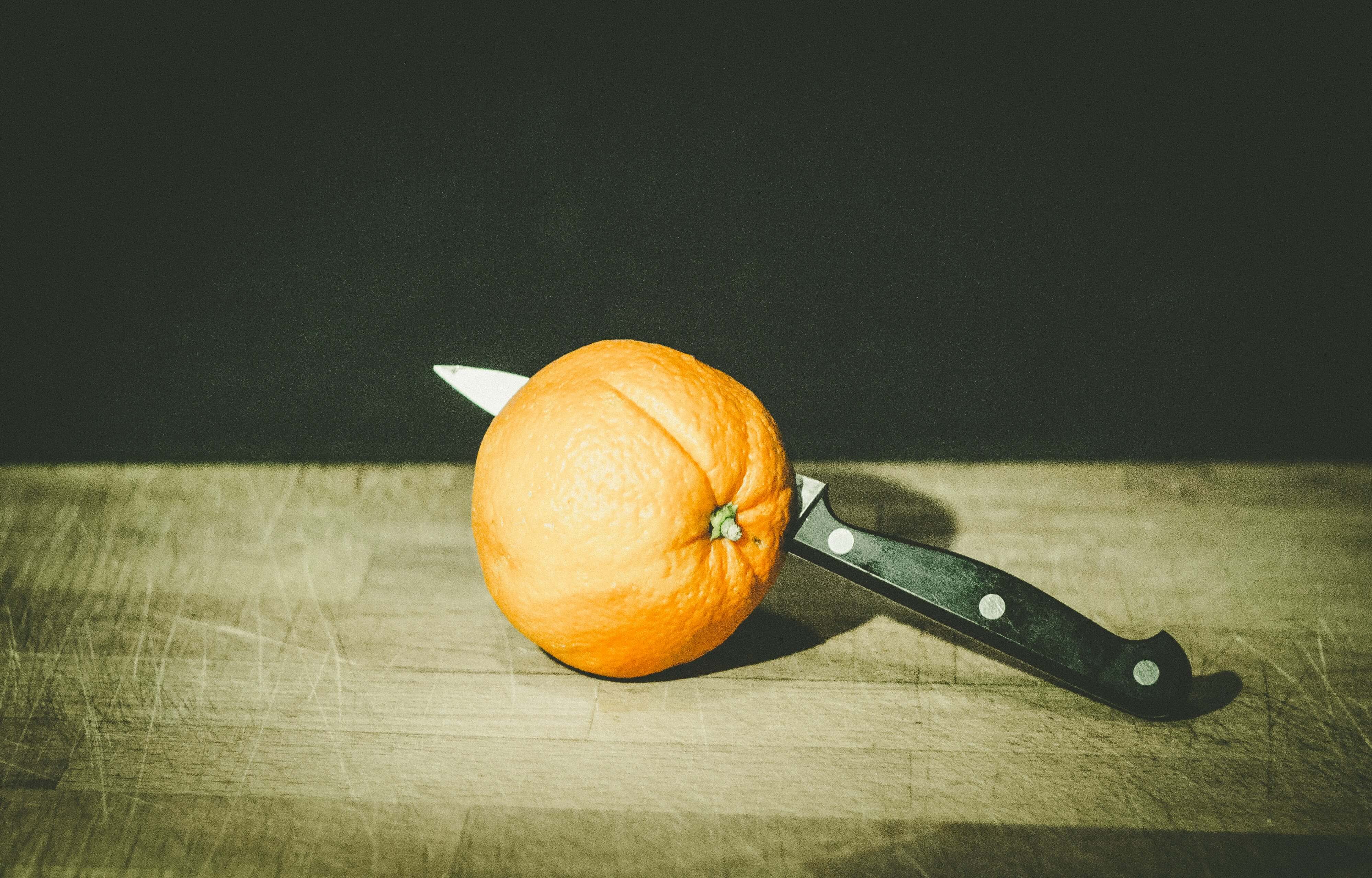 Ein Messer steckt teilweise in einer Orange, die auf einer Holzoberfläche vor einem dunklen Hintergrund liegt.