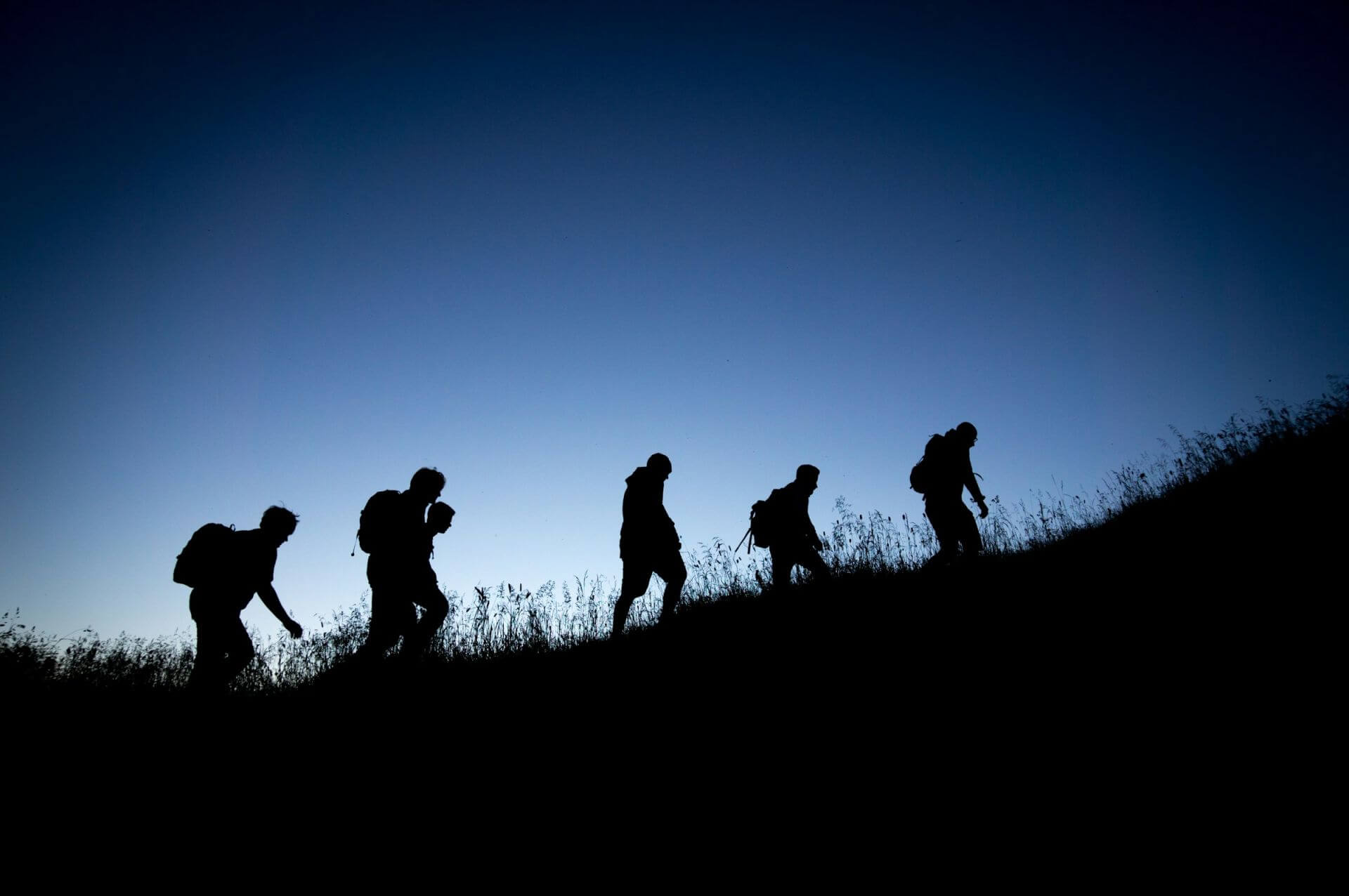 Five people with rucksacks hike uphill at dusk or dawn against a clear blue sky in the silhouette.