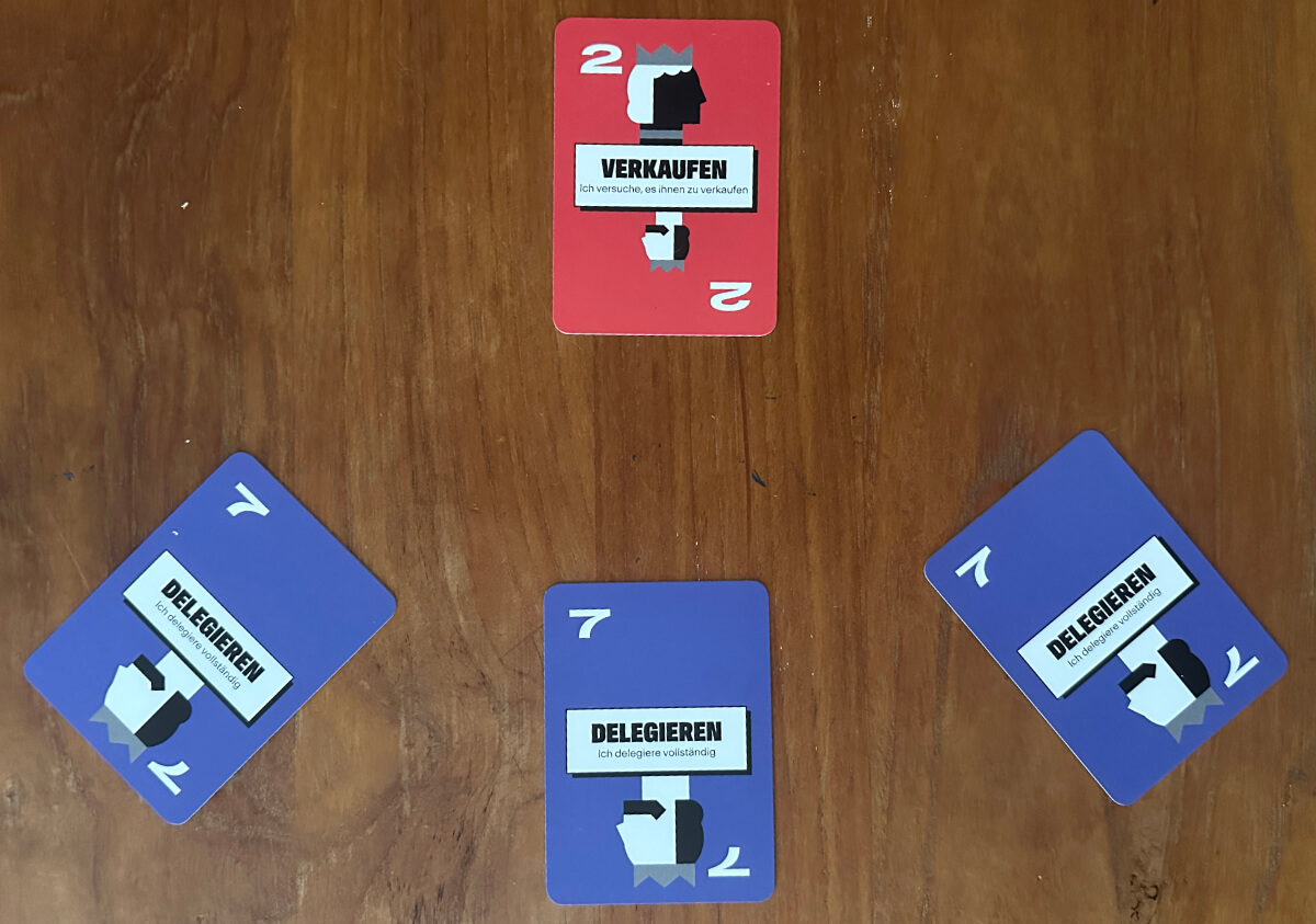 Vier Spielkarten sind auf einer Holzfläche angeordnet: eine rote Karte oben mit der Aufschrift "VERKAUFEN" und drei blaue Karten mit der Aufschrift "DELEGIEREN" unten und an den Seiten.