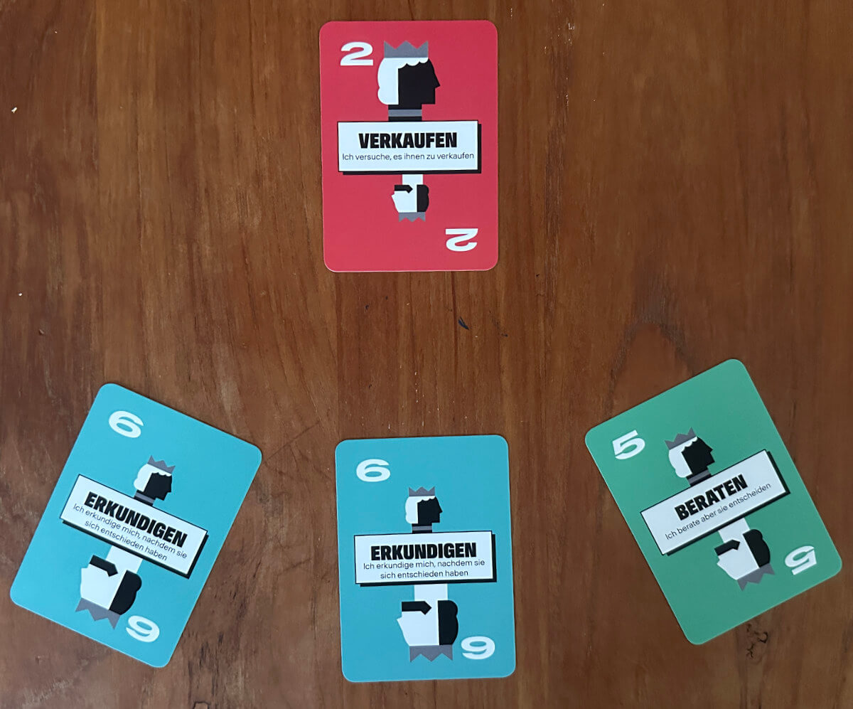 Auf einem Holztisch sind vier Spielkarten ausgelegt: oben eine rote Karte "Verkaufen", darunter drei Karten mit der Aufschrift "Erkundigen" in Blau und "Beraten" in Grün.
