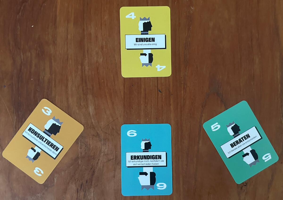 Vier Spielkarten in deutscher Sprache sind auf einer Holzfläche ausgelegt. Die Karten sind mit 3, 4, 5 und 6 nummeriert und zeigen verschiedene Wörter und Symbole.